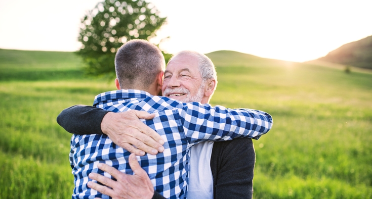 An adult man embraces his father while they stand in a field with the sun shining behind them.