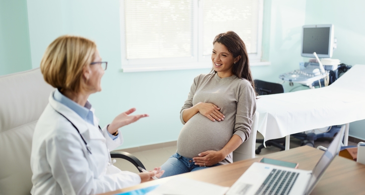 Pregnant woman smiles and holds her belly as she speaks with her clinician in a doctor's office. 