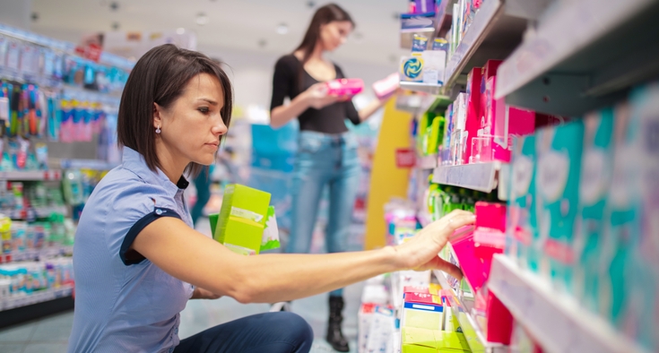 A female customer wearing a short-sleeved top and pants kneels down in the feminine care aisle of a store and selects boxes of menstrual care products to buy. 