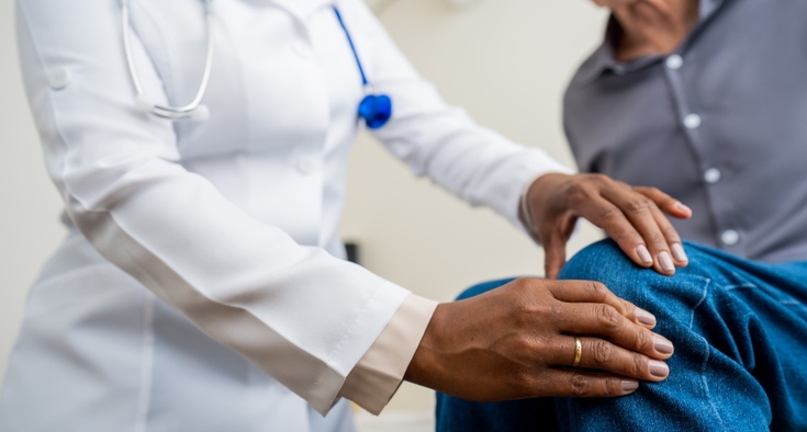 A doctor in a white coat examines the knee of a male patient wearing blue jeans. 