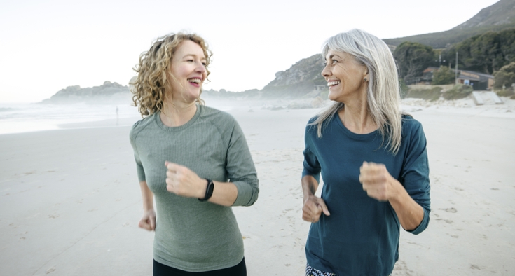 Two middle-aged women smile and talk as they walk side-by-side on a beach.
