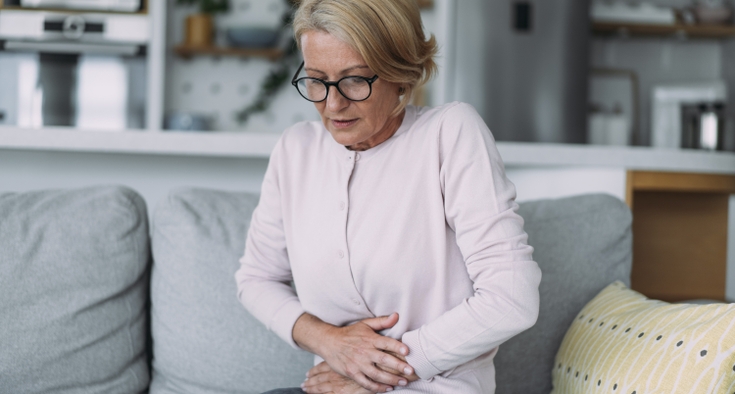 A woman sits on a couch and holds her pelvis in discomfort.