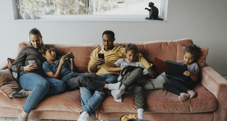 A family of mom, dad, and three kids all sit on a couch using electronic devices.