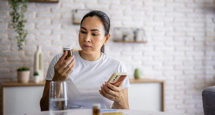 A woman sits at a table. She is holding her smartphone in one hand and reading the label on a prescription bottle in the other hand.