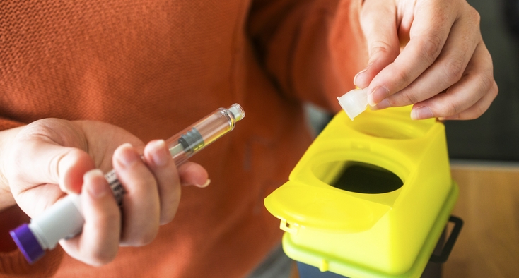 A person holds an autoinjector pen over a sharps safety disposal box.