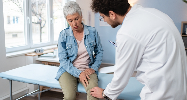 A doctor in a white coat extends his left hand to touch the knee of an elderly woman with short hair who is sitting on an exam table. 