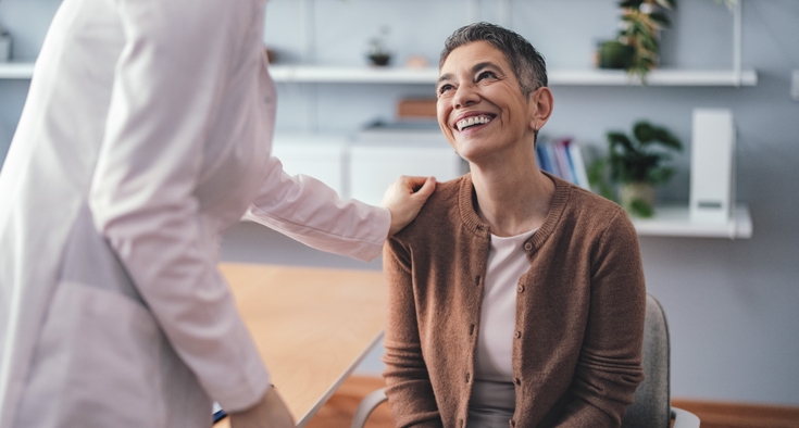 A seated woman smiles as a doctor places a hand on her shoulder.