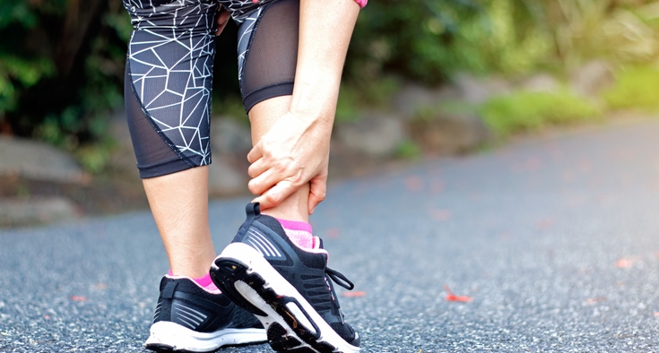 The photo shows a woman from the waist down who is wearing black patterned leggings and black sneakers. She has both feet on pavement and she is bending down to hold her right ankle with her right hand. 