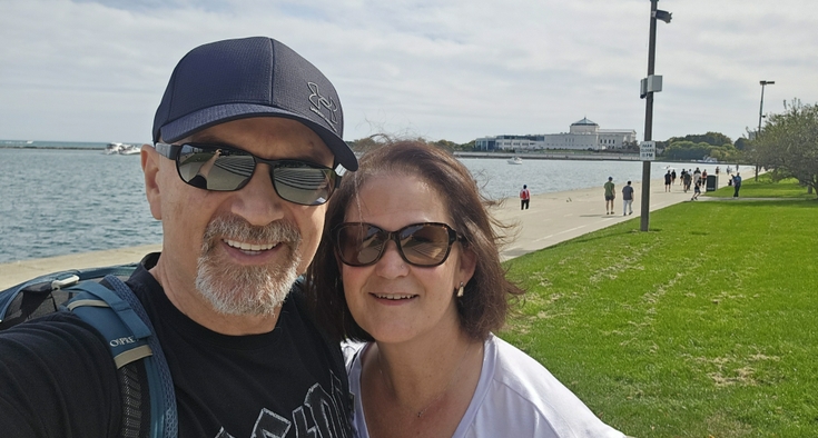 Arben and Michelle Koliqi smile while standing in front of a river on a sunny day.
