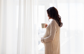 Pregnant woman standing at window holding mug.