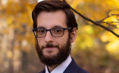 Headshot of Benjamin Suarez-Jimenez Ph.D. He is wearing a white button up shirt and dark blue blazer. He is standing outside during fall in front of a yellow tree. He had neat brown hair, a beard, and is wearing glasses.