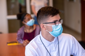 Young adult male wearing a mask listens intently to lecture, while a young woman sits behind him also listening. 