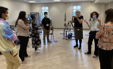 Five students, an interpreter and a physician stand talking in a room with exercise equipment. 