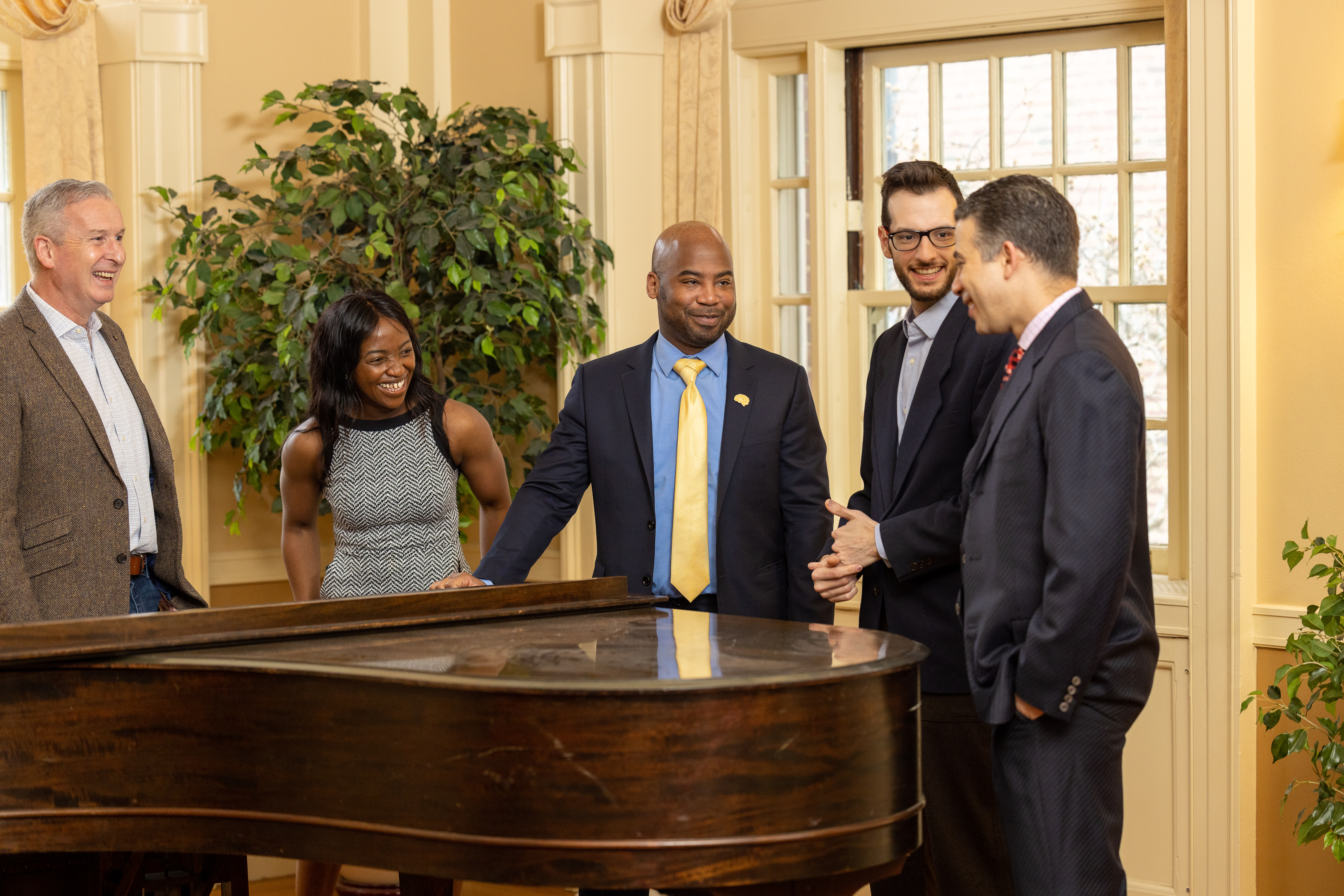 Five people stand talking around a grand piano.