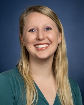 This is a head and shoulders pictuer of Victoria Popov who has blonde hair and blue eye. She is smiling in front of a blue background and wearing a dark green shirt.