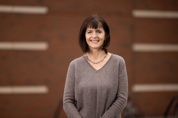 Margot Mayer-Proschel smiling with short brown hair, wearing a tan sweater, standing in front of a brick wall.