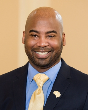 Nathan A Smith is wearing a blazer over a light blue shirt and yellow tie, he is smiling.