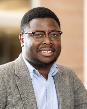 Mark Osabutey smiling while wearing glasses a grey tweed blazer over a light blue collard shirt.