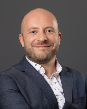 Ian Fiebelkorn smiling in a head and shoulders shot wearing a dark blazer and white button up shirt with collar and small blue pattern.