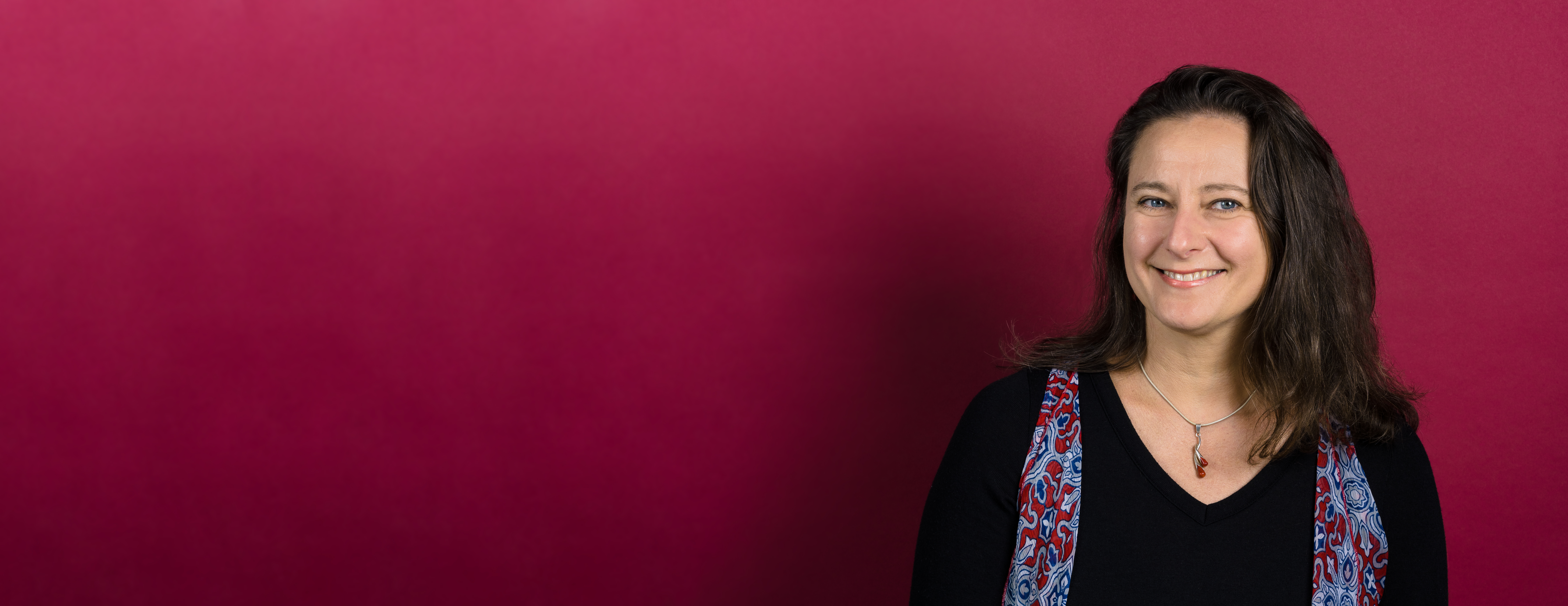 Ania Majewska, PhD, smiling in front of a raseberry colored wall wearing a black top with blue and raseberry scarf.