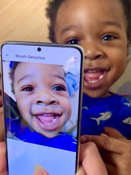 a smartphone is held in front of a smiling baby. The smartphone screen shows a view of the baby's face, with a blue box around the mouth. The top of the screen reads "Mouth Detection".
