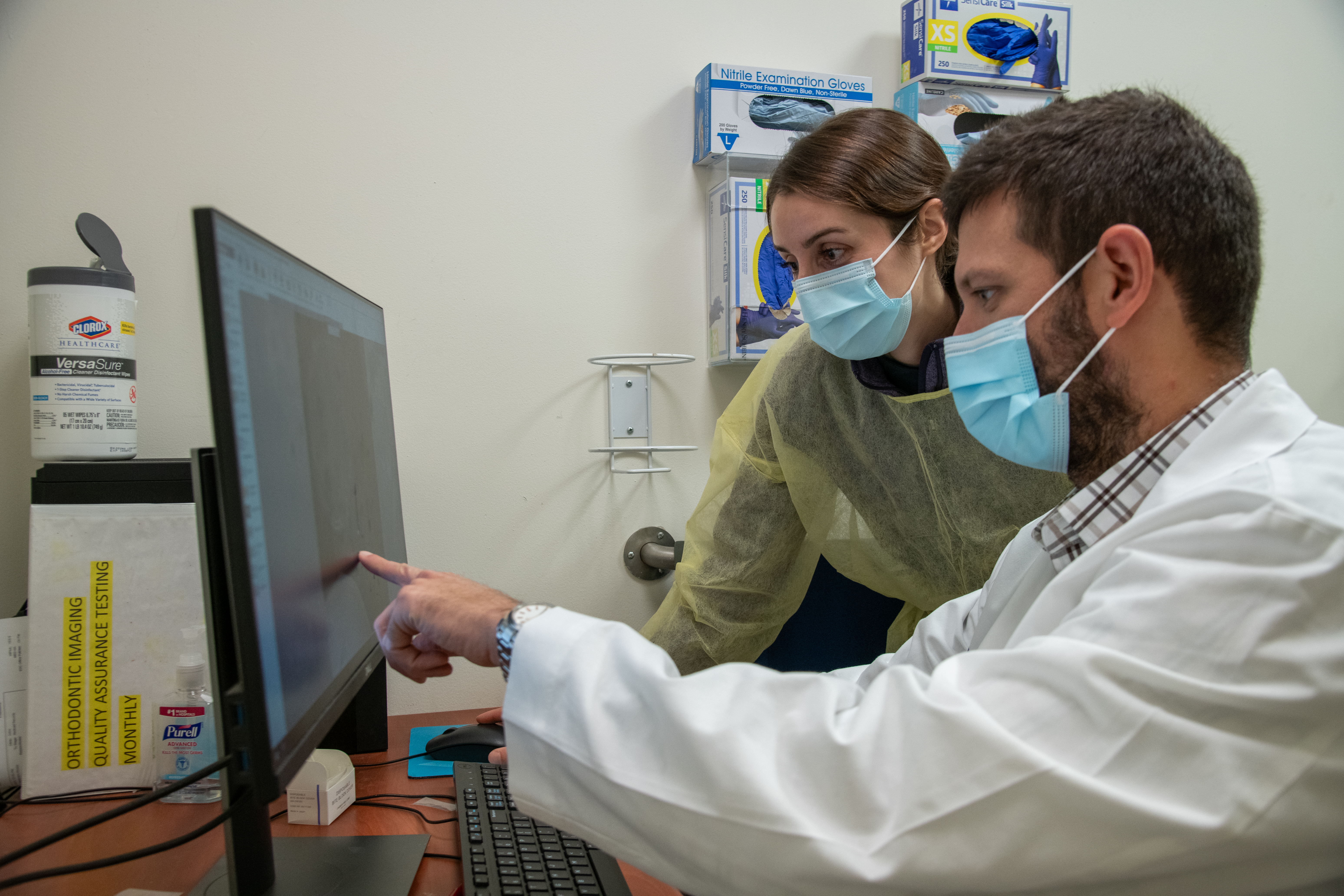 A man in a lab coat points at a computer screen, explaining something to the person next to him
