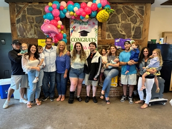 Joe and Karen Ferrari stand in front of a large fireplace, surrounded by their children and young grandchildren. Pink and blue balloons can be seen in the background, as well as a banner with the image of a graduation cap on it, reading "Aloha!" and  "Congrats".