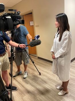 Dr. Xiao stands in front of two cameras, wearing a white lab coat. Two microphones are held in front of her by reporters.