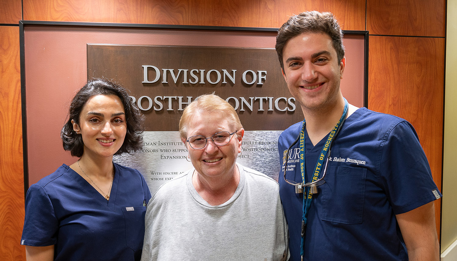 A person in a grey t-shirt stands between two people wearing dark blue scrubs.