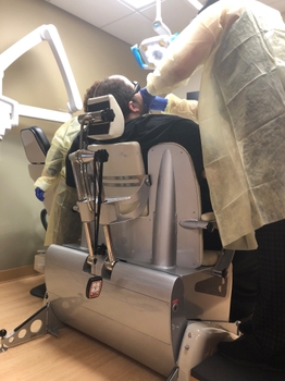 A bariatric patient is seen from behind, seated on a wide dental chair. A dentist examines the patient's mouth from the side.