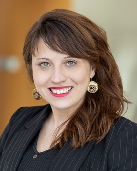 Manoela Fogaca, PhD, is smiling with red lipstick, gold earrings, hazel eyes, and medium length wavy brown hair. She is wearing a black blazer and is standing in front of a brick wall.