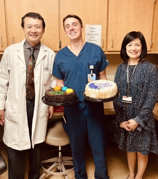 A man in scrubs smiles holding two birthday cakes, between a man in a lab coat and a woman in a dress.