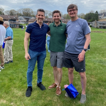 Three men stand outside in the grass. Multiple other people and houses are seen in the background.