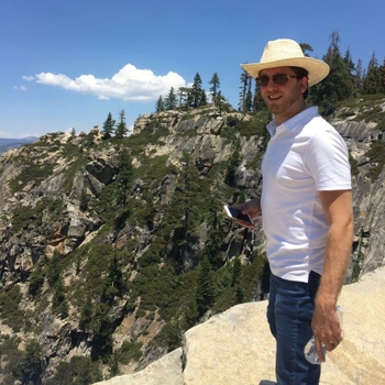A man in sunglasses and a straw hat stands at the edge of a cliff, with a view of surrounding tree-covered rocks.