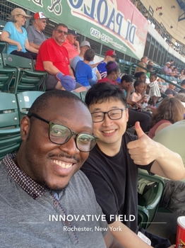 Two men in glasses sit in chairs on the bleachers of a crowded stadium. The man in the back is giving a thumbs-up. Text on the bottom of the picture reads "Innovative Field - Rochester, NY"