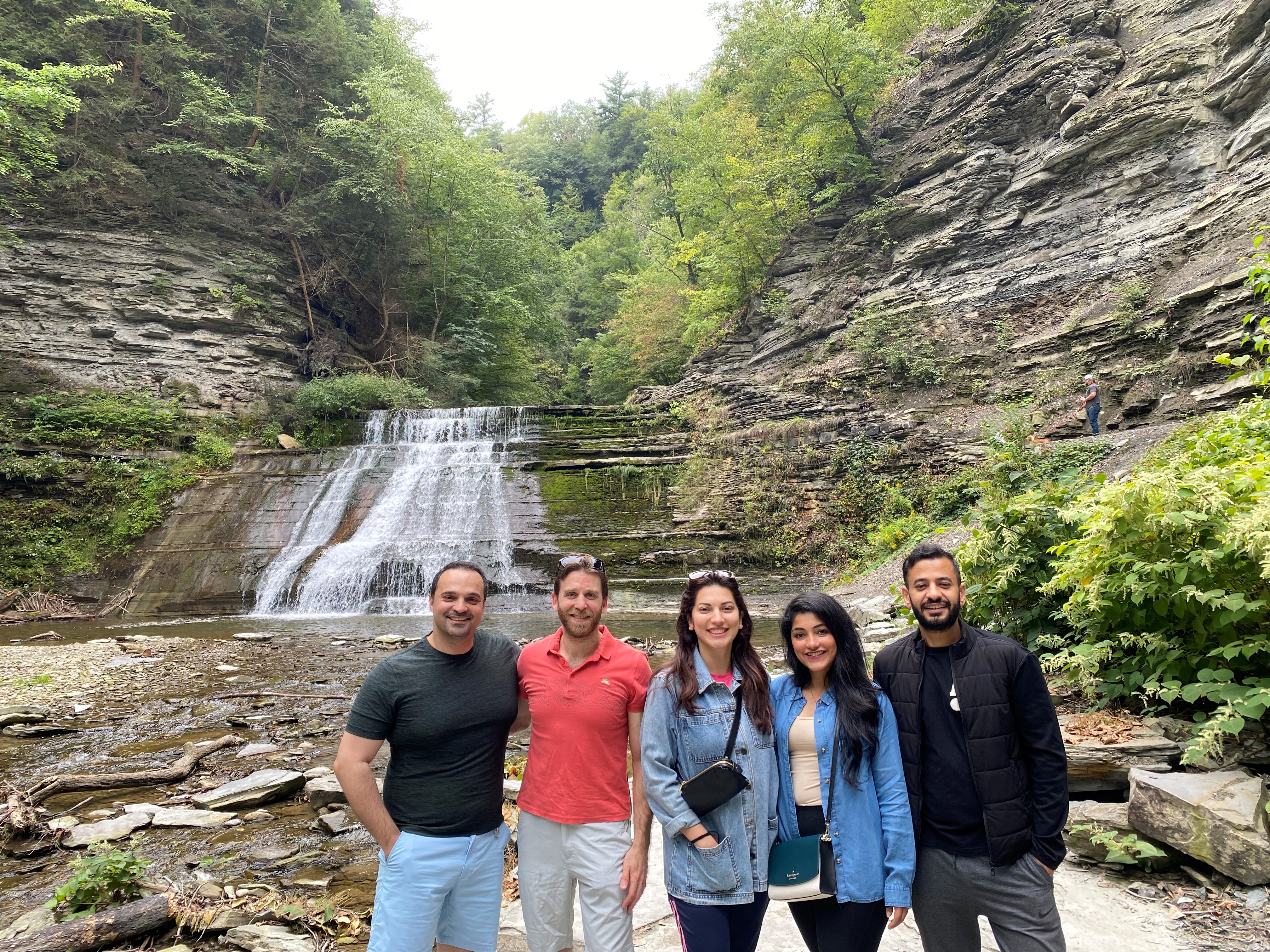 5 people stand in front of a small waterfall. Shale cliffs and many trees are visible in the background.