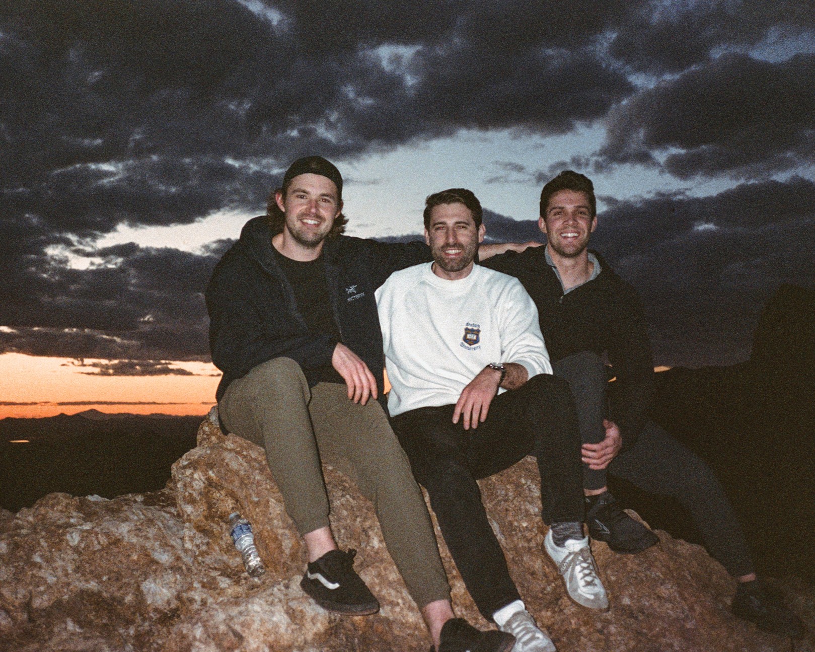 Three men sit on a large rock in front of a sunset.