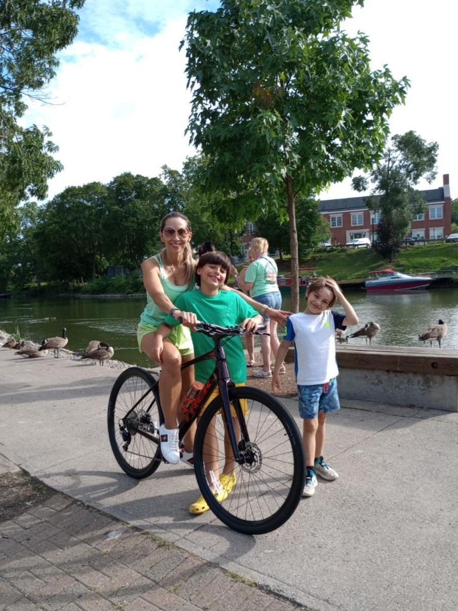 A woman on a bike smiles next to two children. They are next to a canal, with geese behind them.