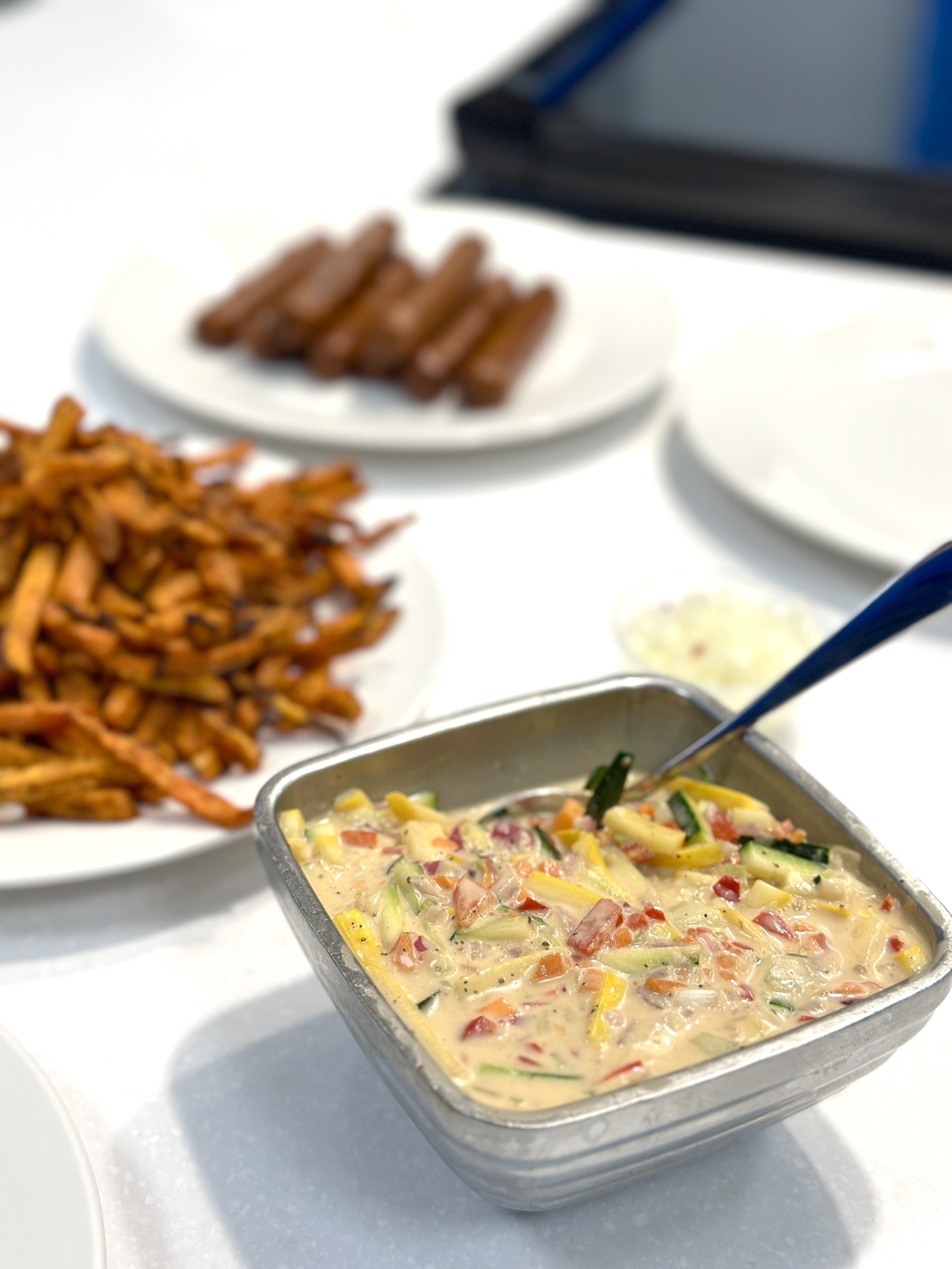A bowl with colorful vegetables mixed with vegan mayonnaise and seasoning sits on the counter in front of a plate of sweet potato fries.