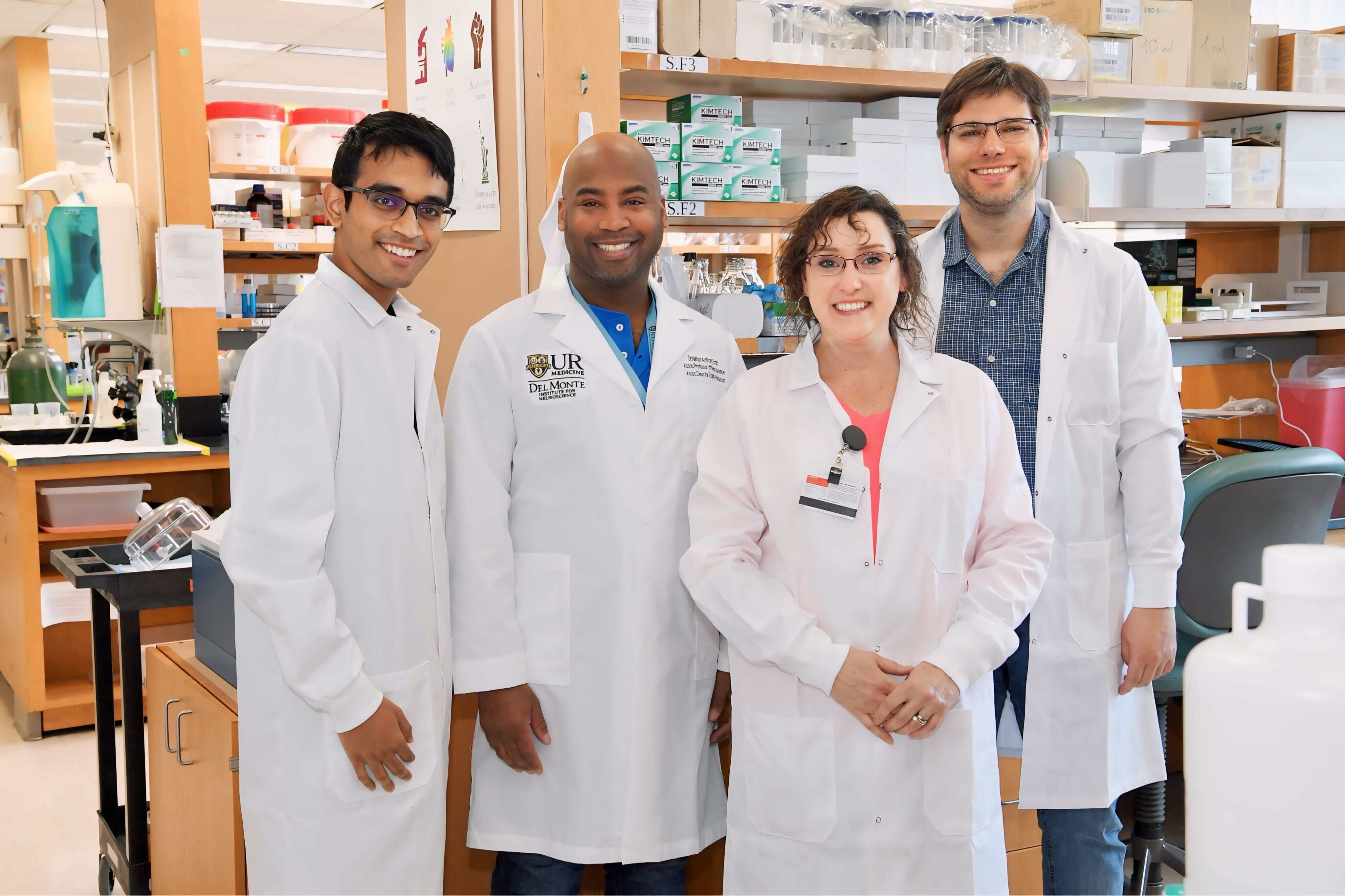 Four people stand together smiling in lab wearing white lab coats.