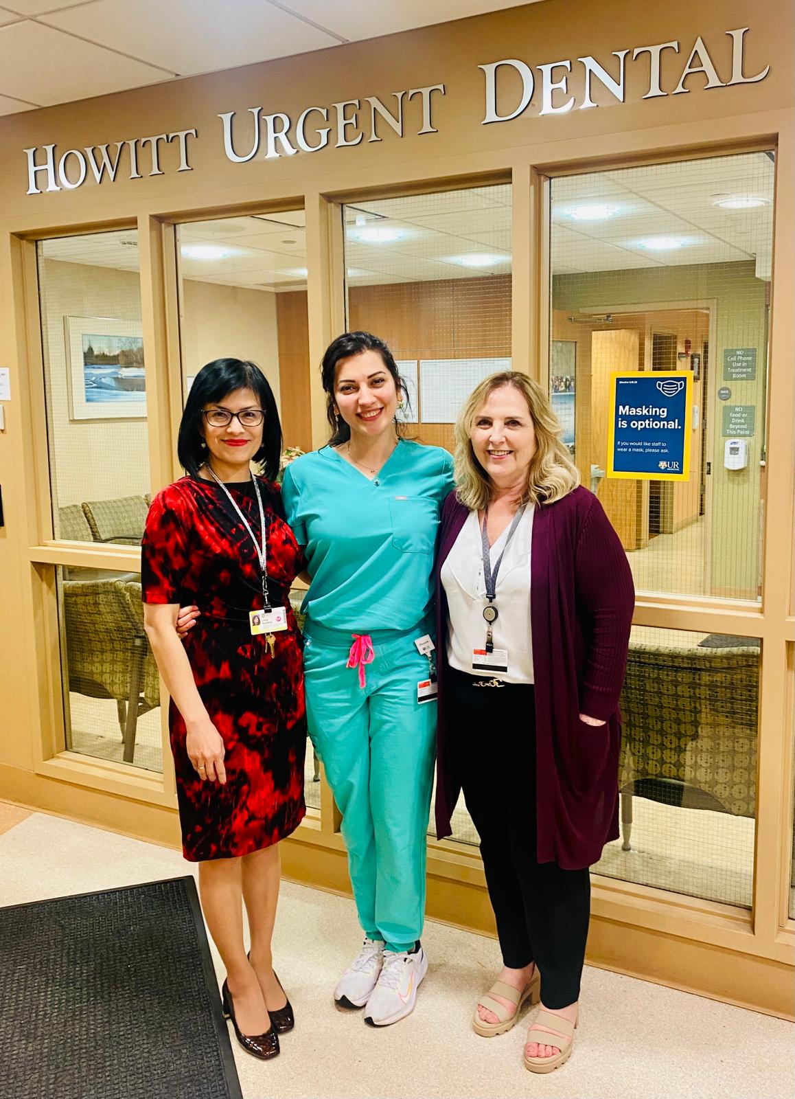 A woman in scrubs stands between two other women in front of Howitt Urgent Dental Care.