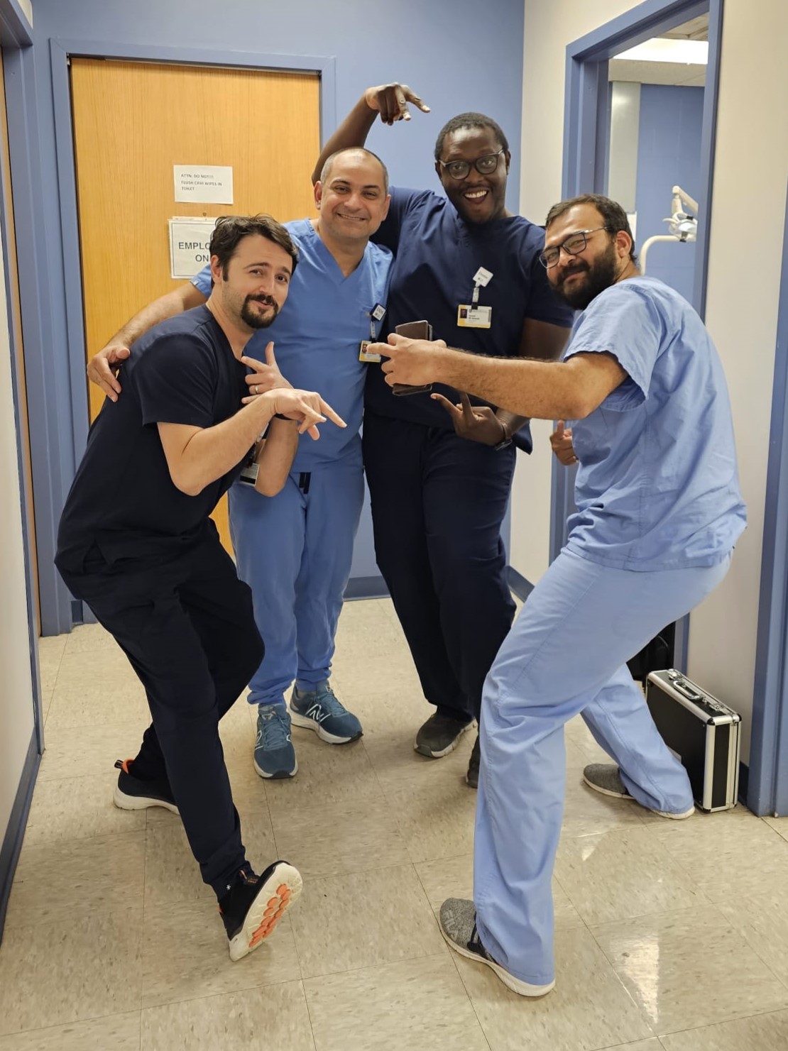Four men in blue scrubs acting silly in a hallway. 