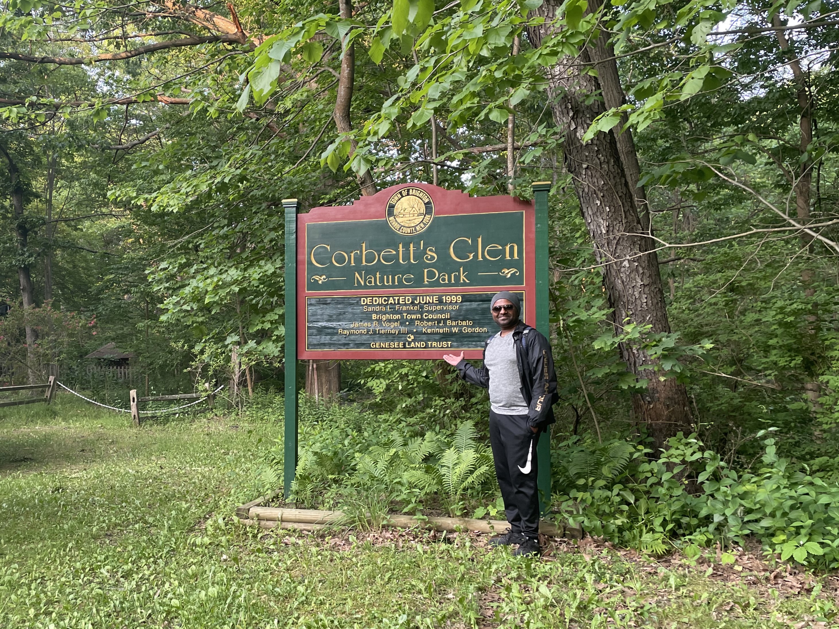 A man stands in front of a park sign that says "Corbett's Glenn Nature Park", surrounded by many green trees and plants.