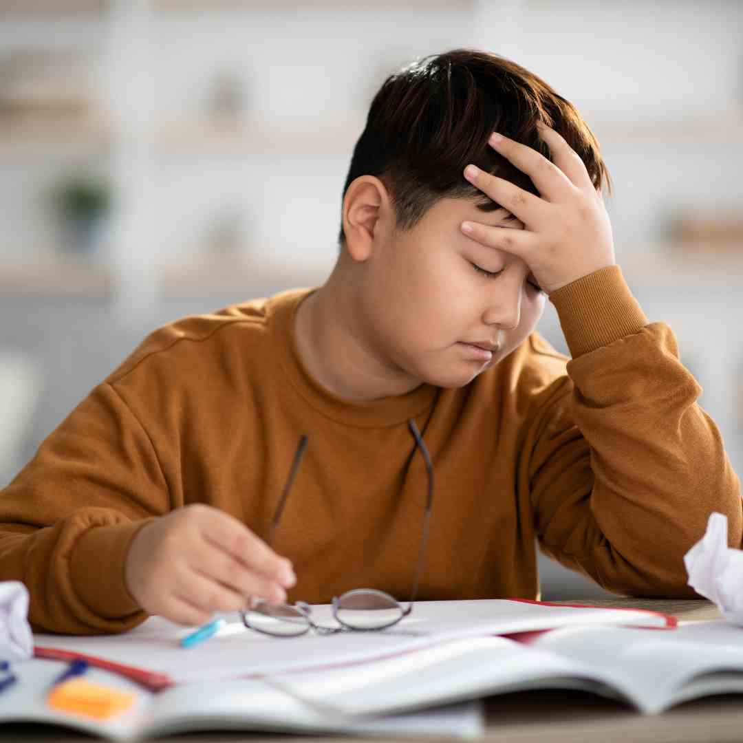 A young boy student holds his head in pain while in class sitting at a desk. He is holding his glasses in his hand.