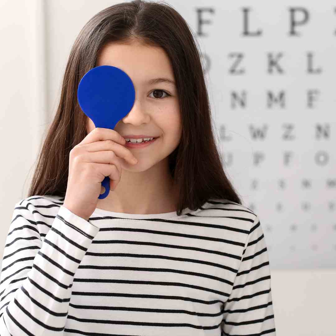 Young girl smiles and holds paddle in front of right eye while looking into the camera. Behind her is a vision exam poster. 