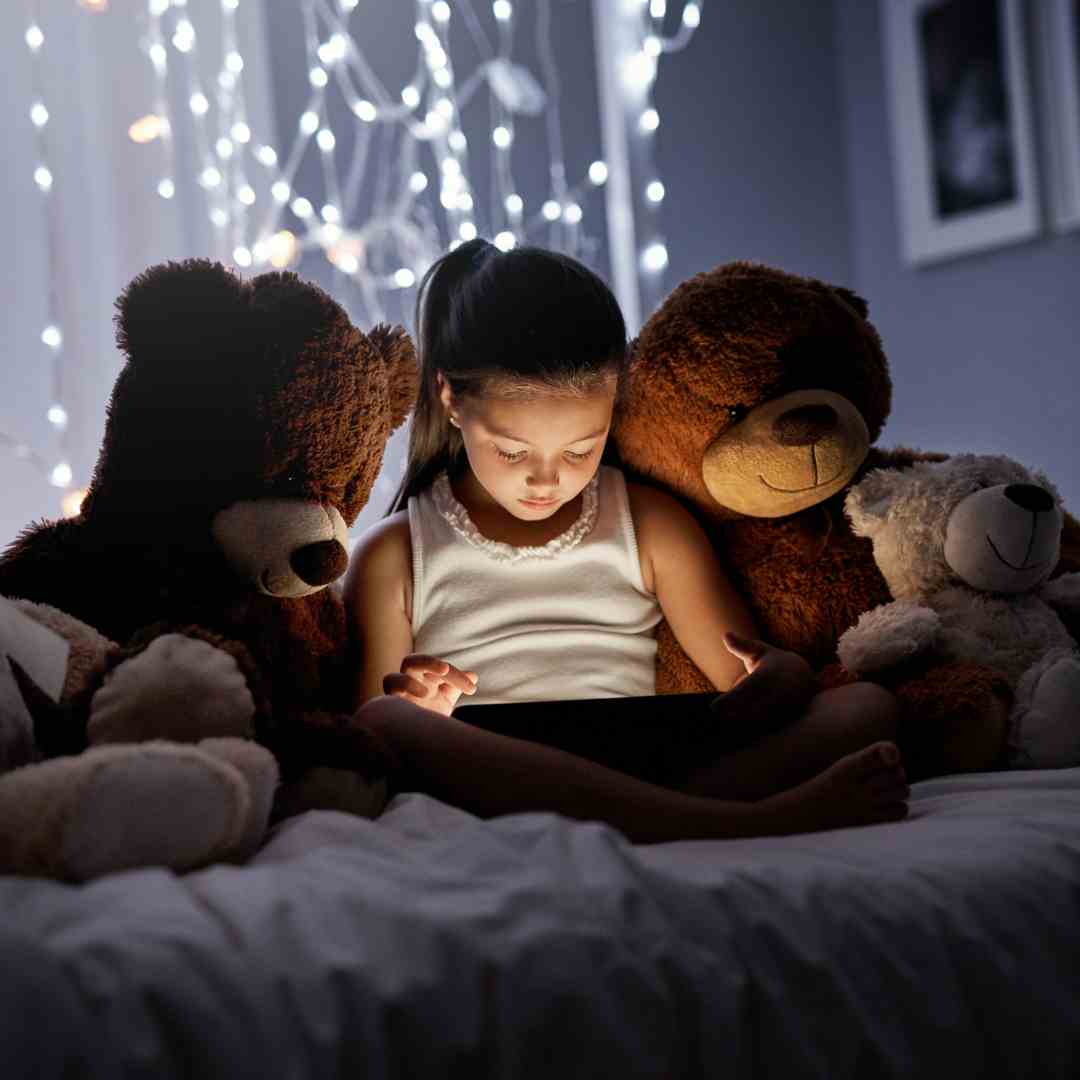 Young girl looking at a bright tablet in the dark with two stuffed teddy bears on each side of her.