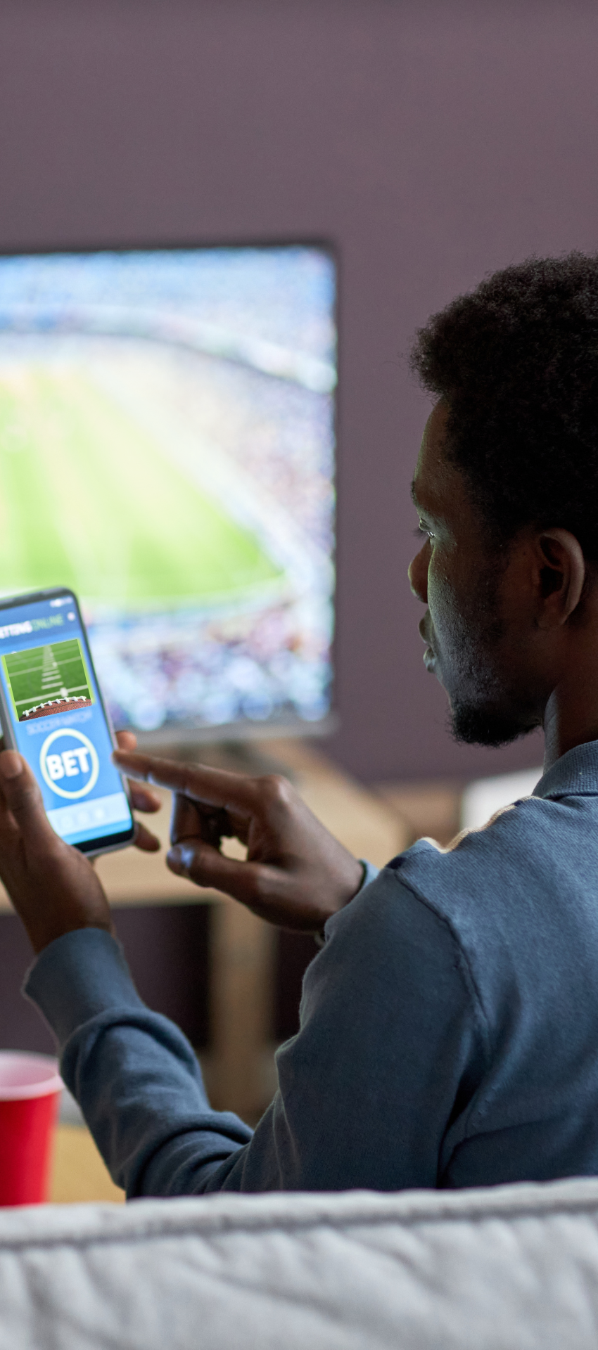 man sits on couch looking at smart phone placing a bet on a sport he is watching.