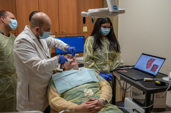 A person in a lab coat uses a small mirror and an intraoral scanner device in a patient's mouth while 3 other people observe.