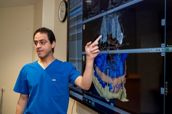 A man in scrubs points to a screen with digital images and 3d models of a jaw and teeth.