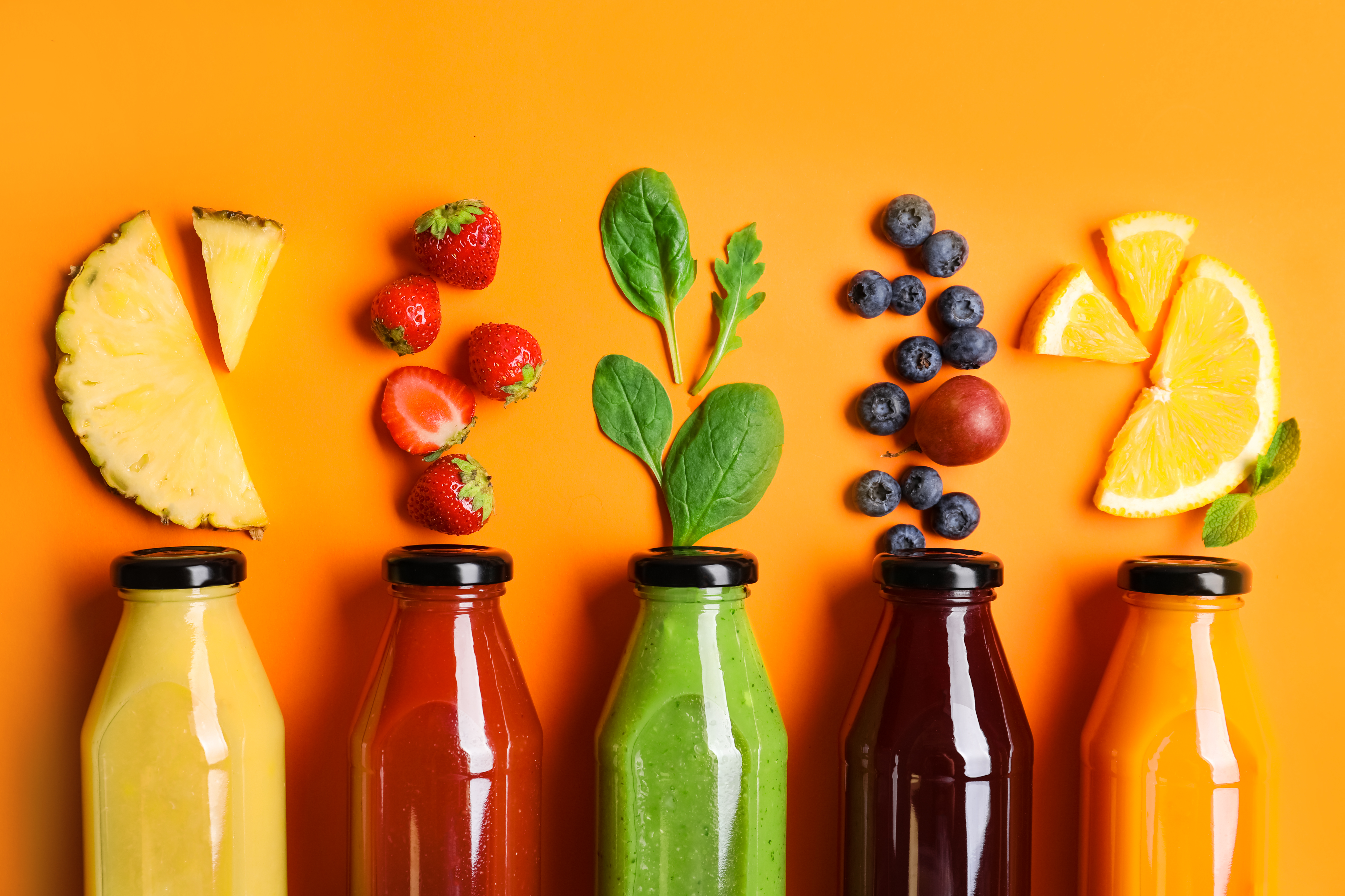 This vibrant image showcases a colorful array of fresh fruit, vegetables, and corresponding juices arranged against a bright orange background. Five glass bottles, each filled with a different colored juice, are neatly lined up in the foreground.
